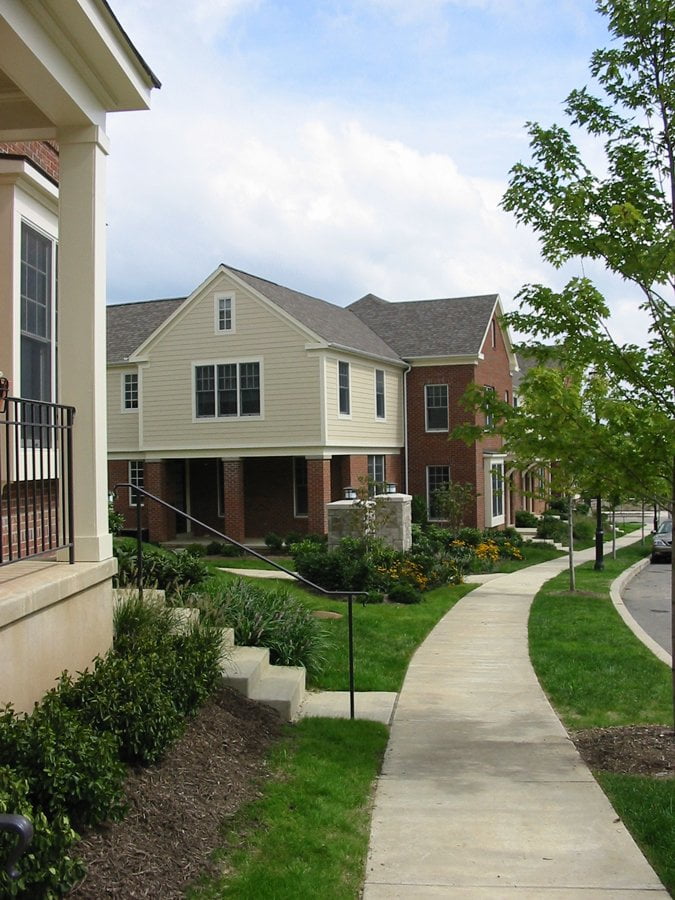 Residential area with a walkway, green grass, trees, and several buildings under a blue sky.